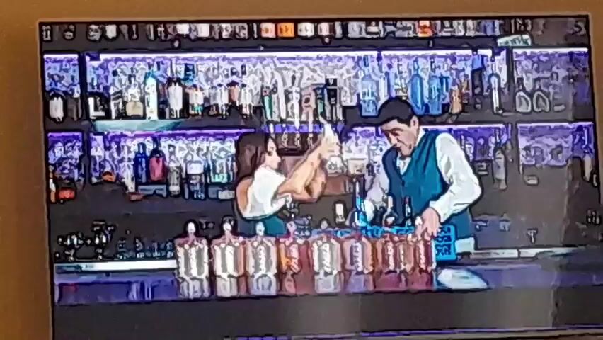 The video displays a short, looping animation, similar to a GIF. The animation features two stylized frames, one with a bar scene and one with a person in a room looking at a laptop. The first frame shows a man and a woman in a bar setting, drawn in a cartoonish style with thick black outlines and vibrant colors, resembling a comic book or a cel-shaded animation. The man, wearing a teal vest over a white shirt, is behind the bar, tending to what appears to be drinks or bottles. The woman, with dark hair, is in front of the bar, facing him, seemingly engaged in conversation. The bar itself is well-stocked with numerous bottles, illuminated by what looks like purple neon lights. There are also several rows of identical, clear bottles or glasses on the counter in the foreground. The second frame shows a person, mostly from behind, in a dimly lit room. The person is looking at a laptop, which is open on a table. Next to the laptop, there is a glass of water and two trophy-like objects that glow with an orange light. Text appears on the screen: "CHRISTINA YEGOYAN" and "ANJIGYAR YAR". This frame also features a white, wispy, vertical line on the right side of the screen, possibly an artistic effect or a visual anomaly. The animation quickly alternates between these two frames, giving the impression of a video clip with a specific style, possibly for a music video or a title sequence.