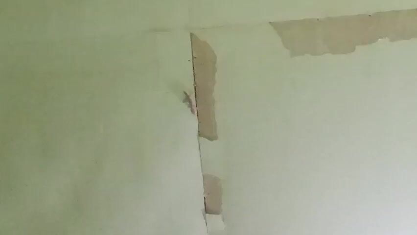 A video records a section of a wall, which is painted light green and shows signs of damage and repair. A vertical seam or crack runs down the center of the frame, with areas of the wall on either side appearing lighter in color, indicating patching or primer. The texture of the wall suggests it may be drywall or plaster. The lighting is dim, and no individuals or other objects are visible in the frame. The video is static, with no movement or camera motion.