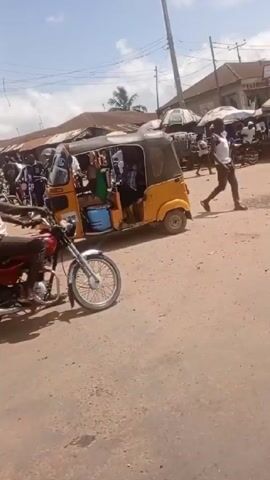 The video shows a street scene in what appears to be a developing country, possibly Africa. Several yellow auto-rickshaws, also known as "keke" or "tuk-tuks," are visible, with passengers inside. One person wears a blue shirt with a black jacket, holding a book. The rickshaws are parked near a street with other people, motorcycles, and open-air stalls.  A beat-up white minibus is also visible, parked close to the rickshaws, with faded paint and a damaged front bumper. Passengers are visible inside. The overall impression is that of a bustling transportation hub in a busy urban area.