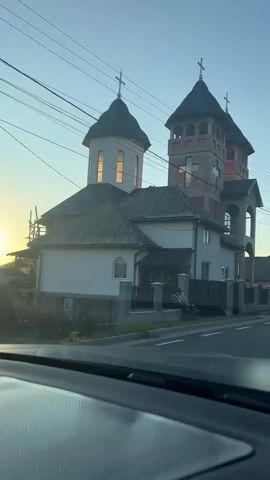 The video shows a partially constructed building with church-like architecture, featuring multiple towers topped with crosses. The building is made of exposed brick and concrete. It is situated next to a smaller, completed house with a fence. Power lines are visible above the structures, and the sky suggests either sunrise or sunset. The camera appears to be inside a vehicle, looking out through the windshield, as indicated by the dashboard in the foreground and the movement suggesting the vehicle is in motion.