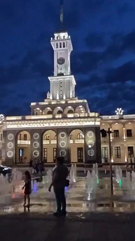 This video shows a night scene outside a building with a prominent clock tower. The building is lit with bright lights, and there are several arched windows on the lower level. In the foreground, there is a plaza with a fountain. The fountain has colorful lights, and the water is spraying in various patterns. Two people, an adult and a child, are standing near the fountain. The adult is facing away from the camera, and the child is facing the fountain. The sky is dark blue with some clouds. The video is shot from a low angle, looking up at the building.