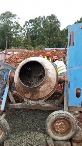 The video shows a construction site where a cement mixer is being operated. A man wearing a blue hard hat and a yellow safety vest is standing next to the mixer, working a hand crank attached to the machine. The mixer is partially filled with rocks and what appears to be concrete.  In the background, other workers wearing red hard hats and yellow vests are visible near a pile of reddish-brown dirt. The ground around the mixer is covered in small gray gravel. The weather appears overcast, and lush green trees fill the horizon. A partial view of a building and utility poles can be seen in the background.