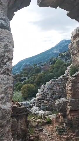The video shows a view of an ancient stone structure, possibly a ruin. The scene is shot from within an arched opening, framing a view of a tree-covered hillside and a distant mountain range under a cloudy sky. The arch and surrounding walls are made of irregularly shaped stones, with some sections revealing brickwork. The camera pans upwards, showcasing the stone construction and the open sky above.