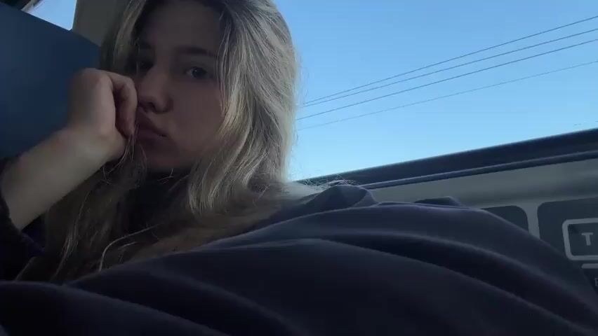 This is a close-up, first-person perspective video of a young woman with blonde hair. Her left hand is supporting her chin, and she is looking towards the camera with a pensive expression. She is wearing a dark hooded sweatshirt. The background shows a blue sky with power lines and a window frame, suggesting she might be traveling on a bus or train. The video consists of only a few seconds of footage, during which her facial expression shifts slightly, and her eyes close and then reopen.
