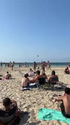 The video shows a beach scene under a clear, sunny sky. A group of shirtless young men are playing with a soccer ball near the water's edge. Some are actively participating in keeping the ball in the air, while others stand nearby. In the foreground, several people are sitting in beach chairs or on towels, observing the game. The sand is light-colored, and the ocean stretches out to the horizon, with a few swimmers visible in the distance.