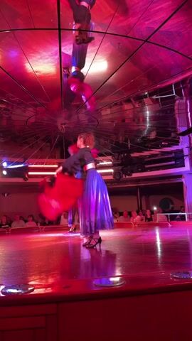 A female dancer on a stage, illuminated by red and pink lights. She is wearing a long, flowing blue and purple dress and holding a red scarf. She is spinning and twirling the scarf around herself. There is another dancer behind her, also in a dark outfit. The ceiling of the room is mirrored, reflecting the dancers and the lights. In the background, an audience is seated, watching the performance. The stage is wooden and appears to be made of a reddish-brown material. The overall atmosphere is theatrical and vibrant.
