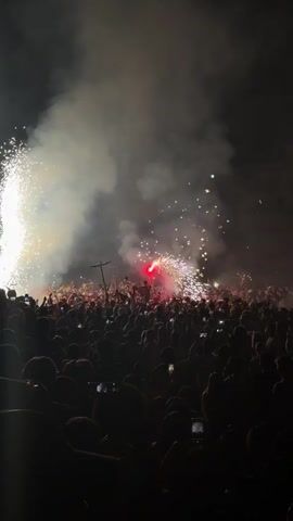 The video shows a dark night scene filled with a large crowd of people. Fireworks are exploding in the background, creating bright flashes of light and smoke. Many people in the crowd are holding up their phones, presumably to record the event. The atmosphere appears to be festive and exciting.