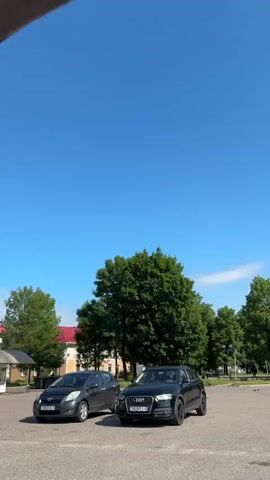The video captures an outdoor scene on a clear, sunny day. The sky is a vibrant blue, with a few wispy clouds visible. In the foreground, there is a paved area, likely a parking lot or plaza. On the left side of the frame, two cars are parked: a dark-colored hatchback and a black SUV. Further back, a line of lush green trees forms a backdrop. Beyond the trees, several buildings are visible, including what appears to be a church with a white facade and a green steeple topped with a cross. A few people can be seen scattered throughout the scene, some walking and others standing. The overall atmosphere is calm and tranquil.