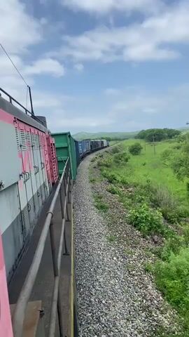 A freight train is moving along a curved track through a green, rural landscape. The camera is positioned on the side of the train, looking forward and slightly to the right. In the foreground, the side of a train car is visible, painted pink and grey with white doors. A metal railing runs along the edge of the train car. The track is covered in gravel. Beyond the track, a vast area of lush green grass and bushes stretches into the distance. Trees dot the landscape, and rolling hills are visible further back. The sky is blue with scattered white clouds. The train appears to be a long string of boxcars, primarily dark blue and green, following the curve of the track. The overall impression is a journey through a natural, open environment.
