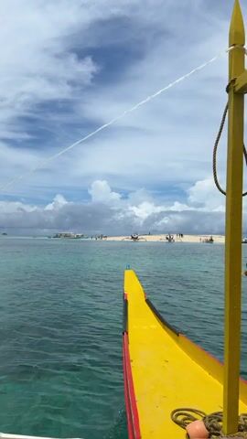 A daytime, eye-level, static shot from a boat approaching a small island. The boat has a yellow bow with a red stripe and a yellow support pole with a hanging rope. The island is a thin strip of sand with several people walking around. There are a few other boats docked near the island. The water is clear and turquoise. The sky is blue with puffy white clouds. The motor of the boat is audible in the background.