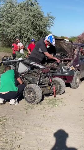 The video shows a group of people gathered around a modified quad bike and a damaged maroon car on a dirt ground. One man in a green shirt is crouching near the quad bike, appearing to work on it, while another man wearing a helmet is reaching under the hood of the car. Several other people are standing nearby observing. The background includes trees, a blue sky, and another modified vehicle with a cage. The overall atmosphere suggests a casual, outdoor setting where people are engaged in tinkering with vehicles.