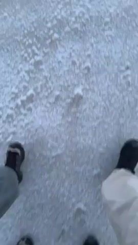 The video shows a first-person perspective of two people walking on a surface covered in ice and snow. The camera is pointed downwards, primarily showing the feet and legs of the individuals as they move. One person is wearing light-colored pants and dark shoes, while the other is wearing darker clothing and footwear. The surface is predominantly white with patches of ice and snow, and a few small twigs are visible on the ground. The movement suggests the people are walking, possibly on a frozen body of water or a snow-covered area.