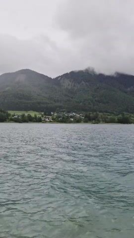 This media file captures a serene scene of a lake with a mountainous backdrop. The water in the foreground is a cool, greenish-blue, rippling gently under an overcast sky. The middle ground shows a lush green shoreline dotted with trees, and further back, a small cluster of buildings, likely a village or hamlet, is nestled against the lower slopes of a large, forested mountain range. The mountains themselves are covered in dense greenery, with some cloud cover obscuring their peaks, suggesting a cool or damp day.

The overall mood is tranquil and somewhat somber, likely due to the cloudy weather. The lighting is diffused, indicating it's either early morning or late afternoon on a cloudy day. There are no people or distinct objects visible in the frame, emphasizing the natural landscape. The setting appears to be Brunn, Austria, a location known for its picturesque lakes and mountainous terrain. The activity, if any, is simply the gentle lapping of the water against the unseen shore or vessel from which the recording is made. No text is visible in the media file.