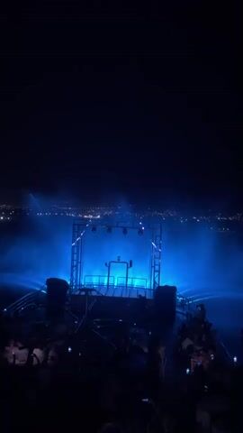 The video shows an outdoor nighttime event with a performance stage in the center, illuminated by blue, yellow, and green lights. Water fountains are also lit with the same colors and are spraying in arcs behind the stage. A crowd of people is visible in the foreground, many holding up their phones, presumably to record the event. The background shows a distant cityscape with lights. The overall atmosphere is festive and lively.