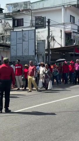 A large group of people, predominantly wearing red shirts and some with red caps, are seen walking along a sunlit street in what appears to be a procession or rally. They move from left to right across the frame. One person near the center carries an umbrella.

The setting is an urban street lined with multi-story buildings, characteristic of a commercial area. Storefronts with signs are visible, including "SALON BLACK Bridal Hair Beauty" and "SALON UNISEX." Further down the street, a circular yellow sign with a spoon and fork icon and the word "FOOD" is prominent, associated with "ALANA FOOD CENTER" written below it in English and Sinhalese script ("අලාරි ෆුඩ්"). A partial menu board to the left advertises "burgers, subs, wrap, hotdogs, pastas, fried rice, cheese kotu, milkshakes, fruit juices" along with a phone number "(30) 350 4473". Red flags or banners are displayed along the street, possibly indicating the nature of the gathering. Overhead power lines crisscross the sky.

The weather is bright and sunny with a clear blue sky and some white clouds, suggesting it's daytime. Shadows are visible on the road surface, cast by the buildings and people. The road itself is asphalt with white lane markings. The overall scene suggests a busy thoroughfare in Delkanda, Sri Lanka, given the visible script and architectural style.