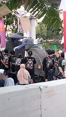This is a vertically oriented video of a group of people gathered outdoors, possibly for an event or ceremony. The background appears to be a somewhat informal setting with some foliage, including large banana leaves on the left side of the frame. In the foreground, a group of individuals, mostly wearing black t-shirts with a floral design, are seen from behind. They appear to be bowing or kneeling in unison, suggesting a religious or respectful observance. Some people are wearing headscarves, including one person in a light brown hijab. In the center of the frame, there is a dark object that resembles a small, vintage scooter or motorcycle, positioned behind the kneeling individuals. A white, textured wall or structure dominates the right side of the frame. There are also some colorful banners or fabric strips visible at the bottom of the frame. The lighting suggests it is daytime, possibly late afternoon or early morning due to the angle of the light and the shadows.