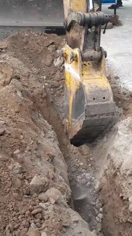 An excavator is digging a trench. The excavator's bucket is submerged in the dirt, and soil is being excavated. The trench is narrow and deep, with dirt piled up on either side. The ground appears to be a mixture of dirt, rocks, and gravel. The scene appears to be an outdoor construction site.