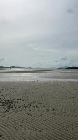 This is a vertical, static shot of a tranquil beach scene. The foreground is dominated by rippled sand, with scattered dark debris. In the mid-ground, shallow water reflects the cloudy sky, and distant islands are visible on the horizon. A few indistinct figures can be seen walking in the distance. The overall impression is one of a quiet, overcast day at the beach.