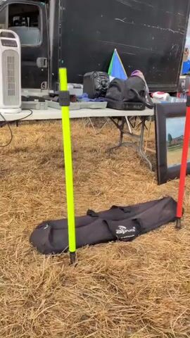The video shows an outdoor market stall with various items displayed on a table and on the ground. Two tall, brightly colored poles, one green and one red, stand upright in the foreground. A black bag lies on the ground between them. Behind the poles, a table is set up with several items, including what appears to be a speaker, a mirror, and other assorted objects. In the background, there is a large black trailer or vehicle, and further back, more stalls and people can be seen, along with a white clock face with Roman numerals. The ground is covered in dry, yellowish grass or straw, suggesting a rural or field setting. The lighting indicates it is daytime, possibly overcast.