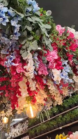 The video features a dense, lush ceiling decoration composed entirely of artificial flowers and greenery. The camera slowly pans across the installation, offering various angles of the vibrant display.

The arrangement includes a variety of faux flora, predominantly orchids in shades of bright pink, deep red, white, and light blue. White hydrangeas are also interspersed throughout. The flowers are complemented by abundant green foliage, including eucalyptus-like leaves and trailing ivy-like plants, creating a rich, layered canopy.

Several exposed, warm-toned light bulbs are integrated within the floral display, casting a gentle glow and adding to the decorative ambiance. The flowers are arranged to hang downwards, creating a beautiful and intricate overhead garden effect. In the background, below the main floral display, shelves with other items, possibly more artificial plants or decor, can be faintly seen. No people are visible, and there is no discernible text.