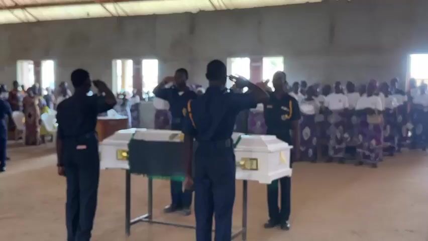 The video shows a solemn ceremony taking place in what appears to be a community hall or a simple indoor venue. In the foreground, four individuals in dark uniforms, possibly a cadet corps or a similar organization, stand at attention and salute. They are positioned in front of two white coffins, each placed on a separate table. The coffins are adorned with gold handles and a dark green cloth draped over them.

In the background, a crowd of people, dressed in a mix of formal and traditional attire, stands or sits, observing the proceedings. Many women are wearing vibrant, patterned wrappers and headscarves. The environment is rustic, with concrete walls and a high ceiling made of corrugated metal, suggesting a simple, functional space. Natural light streams in from windows along the walls. The overall atmosphere conveys a sense of respect and mourning. The audio is predominantly a solemn, slow-tempo musical piece, likely a funeral dirge or a hymn, accompanied by occasional murmuring from the crowd.