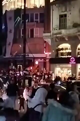 The video shows a crowded street scene at night, with a group of people walking and cars driving. There are several buildings visible, including one with a flag with stars hanging from it. Some of the buildings have lights on, and there are streetlights illuminating the scene. People are walking in the street and crossing between cars. The general atmosphere appears lively and possibly celebratory due to the crowdedness and presence of a flag.
