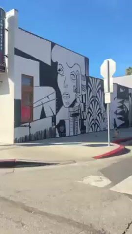 The video shows a view from a moving vehicle driving down a city street on a bright, sunny day.  Buildings with white walls are prominent on either side of the road, some adorned with decorative elements like green awnings, shutters, and a large black-and-white mural of a face. Palm trees are scattered along the sidewalk, casting shadows on the buildings and street. A red car briefly appears, its front wheel visible. The sidewalk has a red curb, and there's a blue lamppost visible at one point. The road surface is typical city asphalt.  The overall impression is that of a sunny day in an urban environment with a mix of architectural styles.