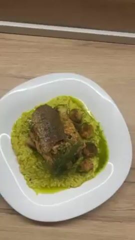 The video shows a single plate of food sitting on a light brown wooden surface. The food consists of a bed of yellow-green risotto, topped with a piece of fish with crispy skin, several roasted brussel sprouts, mushrooms, and a sprig of dill. A bright green sauce is drizzled around the food. The plate is white and has an irregular, wavy edge. The camera slowly rotates around the plate.