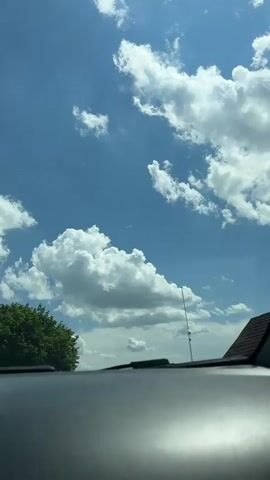 This is a POV video from inside a vehicle looking out at the sky. The sky is blue with scattered white cumulus clouds. A green tree is visible on the left side of the frame. The dashboard of the vehicle is in the foreground, with a windshield wiper visible. The camera is moving forward, indicated by the slight shift in perspective and the blur of distant clouds.