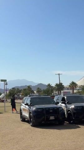 The video opens with a static shot of two black and white Ford Explorer police SUVs parked side-by-side on a dry, sandy field under a clear blue sky. Mountains are visible in the distant background, flanked by palm trees and what appear to be structures for an outdoor event. A street sign for "AVE 100TH ST" is partially visible on the left.

The two police vehicles display "SHERIFF" on their sides and have California license plates "714187" and "711778" respectively. A person in a black t-shirt is seen standing near the left SUV.

As the video progresses, a smiling woman with long hair, wearing a white bandana, a white halter top, and red polka-dot shorts, walks into the frame from behind the police cars. She waves cheerfully at the camera as she passes by, continuing to walk through the event area. The bright sunshine and clear weather suggest a warm, daytime setting, likely in Indio, United States of America.