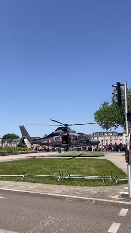 A dark military helicopter with "MARINE" written on its side and a red insignia on its tail hovers just above a grassy area. The helicopter has its door open and appears to be a demonstration or event, as a crowd of people is visible in the background, lining up behind a low barrier. The helicopter is positioned in front of a row of light-colored buildings with a blue sky overhead. In the foreground, a paved surface and a traffic light pole are visible. The scene suggests an outdoor event possibly involving an air show or military display.