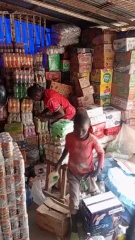 Two young boys are in a small, crammed shop. The shop is filled with various items stacked high, including soda bottles, juice boxes, and snacks. One boy, wearing a red shirt, is standing on a small wooden stool, reaching for items on a shelf. The other boy, in a peach-colored shirt and grey pants, is standing on a box, holding a roll of tape. The environment appears to be a bustling marketplace or small retail space.