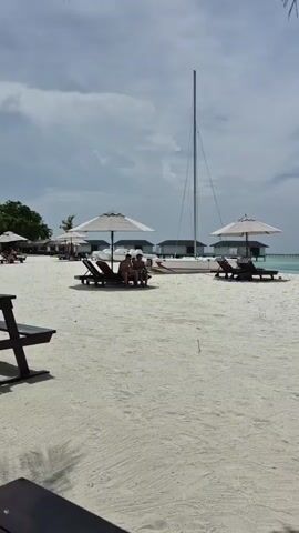 The video shows a tropical beach with fine white sand.  In the distance, there are several bungalows situated over the water, accessible by walkways.  A sailboat is resting on the beach between two sets of beach chairs and umbrellas.  People are relaxing in the chairs, and some vegetation is visible on the left. A wooden bench is situated in the foreground to the left. The sky is overcast, with a mix of gray clouds.