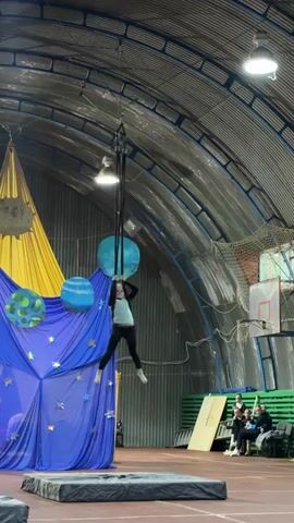 The video captures a young girl performing aerial silks in a gymnasium. She is suspended from a black fabric, moving gracefully through the air with her body in various poses. The setting appears to be an indoor sports facility, with a high, arched, corrugated metal ceiling and exposed lighting fixtures. 

A backdrop is set up to the left of the performer, featuring a large yellow and blue fabric arrangement decorated with celestial elements like planets and stars. One of the planets is depicted with green continents, and another is blue. To the right of the performer, a basketball hoop and backboard are visible, along with bleachers where several spectators are seated, watching the performance.

The lighting in the gymnasium is artificial, casting bright spots from the overhead lights. The floor appears to be a reddish-brown track, typical of a sports venue, with a large, dark mat positioned beneath the aerial silks for safety. The overall atmosphere suggests a performance or competition taking place, possibly a school event or a circus-style exhibition. The girl's focus and the spectators' attention indicate an engaging performance. There is no visible text within the video frame.