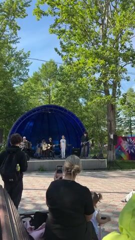 A live music performance is taking place outdoors on a stage under a blue semicircular canopy. There are four musicians on stage. One is playing an accordion, another is behind a drum set, and two are playing guitars. A young boy in a white shirt is standing at the front of the stage. A crowd of people is watching the performance from a park area with trees and a paved walkway. Some people are sitting on a bench in the foreground, and one person is recording the performance with their phone. The weather appears to be sunny with a clear blue sky.