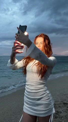 A young redheaded woman is standing on a beach, holding up a phone, and using its light to illuminate her face as she takes a selfie. She wears a white, somewhat sheer dress with long sleeves. The sky is cloudy, with hints of sunset coloring the horizon. The ocean is visible in the background, with small waves lapping onto the sandy beach.