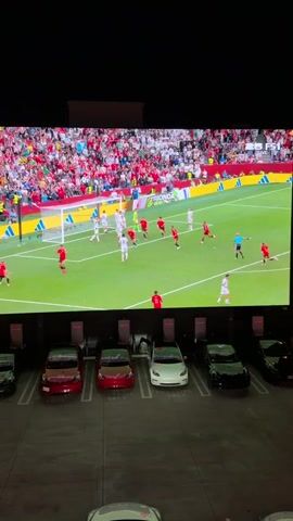 The media file shows a large screen displaying a soccer (football) match. The screen is positioned above a row of parked electric vehicles, suggesting a drive-in or outdoor viewing experience. The soccer field is green, with players in red and white/light-colored uniforms. The stadium appears to be filled with spectators wearing red. The scoreboard visible on the screen indicates "GROUP G NEW ZEALAND 0 BELGIUM 1 HALFTIME". The time of day is likely evening or night, as the sky above the screen is dark and there are lights visible within the stadium. The location context of West Hollywood, United States of America, places this event in a specific urban environment. The overall scene depicts a communal viewing of a sporting event, likely on a large outdoor screen, with the cars serving as viewing booths. The emotions conveyed are focused on the game itself, with the scoreboard indicating a close match at halftime.