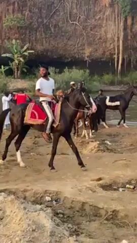 The video shows a group of people near a river, some of whom are riding horses. The horses are dark in color and adorned with decorative saddles. Riders are dressed casually, with one wearing a white shirt and striped pants. Several people are standing around observing the horses and riders. The riverbank has exposed soil and some litter. The backdrop includes a rock face with some vegetation. A small waterfall is visible in the distance.