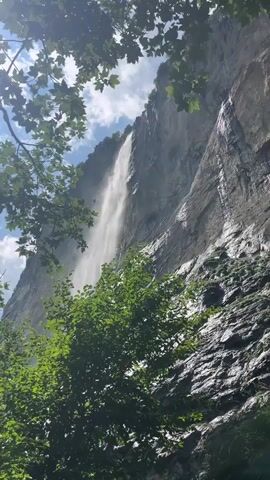A tall, narrow waterfall cascades down a sheer, rocky cliff face. Lush green foliage frames the bottom and left side of the scene, with sunlight dappling through the leaves. The sky above is blue with scattered white clouds. The water is a bright white as it falls, creating a misty spray at its base. The rock face is dark and wet, with streaks of water running down it in places. The overall impression is one of natural beauty and power.
