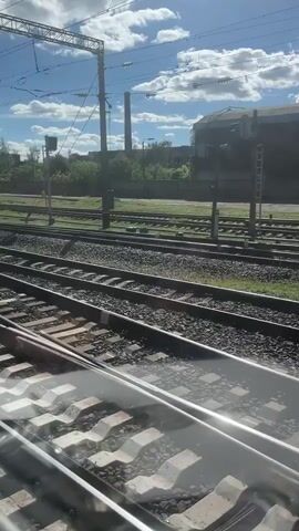 The video shows a railway crossing with multiple sets of tracks. In the foreground, we see the metal rails and concrete sleepers of the tracks, reflecting sunlight. In the middle ground, a paved road crosses the railway tracks, and a person on a bicycle is seen riding across the tracks from left to right. The person is carrying a basket on the front of their bicycle. In the background, there are more railway tracks, power lines with overhead wires, and industrial buildings. The sky is blue with some white clouds. The overall scene is a functional industrial area with active railway lines.