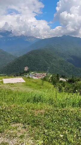 The video opens with a panoramic view of lush green mountains under a cloudy sky. The camera pans across the landscape, revealing a building complex nestled in a valley and a cable car ascending a slope. In the foreground, a grassy hill slopes down to a paved area. As the camera continues to move, it focuses on a group of people gathered near some colorful structures, including a red archway and blue tents. A white van is parked nearby. The camera then pans back to the mountainous scenery.