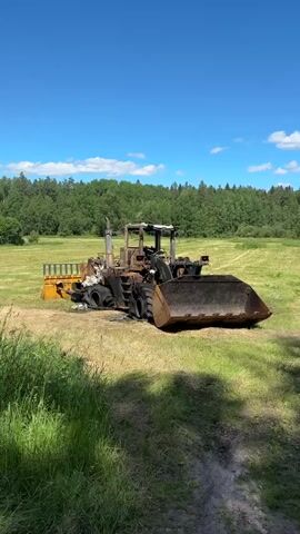 A burnt-out front-end loader sits in a grassy field under a bright blue sky with white clouds. The machine is heavily damaged, with its cab and tires blackened by fire. The bucket of the loader is filled with dirt or debris. The surrounding area is covered in green grass, with a line of trees in the background. The video appears to be a still shot or a slow pan of the damaged vehicle.