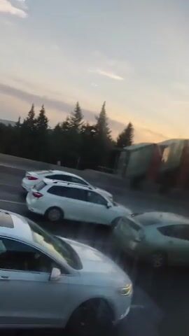The video shows a street scene with cars driving. The camera appears to be inside a moving vehicle, possibly a car, and it pans across the road. In the foreground, the front of a white car is visible. Further ahead, there are several other cars on the road, including a white station wagon, a light green sedan, and a white car with a distinctive rear design. A modern-looking building with red and glass elements is visible on the right side of the road. Trees and a hill are in the background. The sky suggests it is either sunrise or sunset, with soft lighting and some clouds. There are no clear views of people's faces or specific interactions between them, as the focus is on the vehicles and the general environment. The overall impression is of traffic moving along a highway or a busy street.