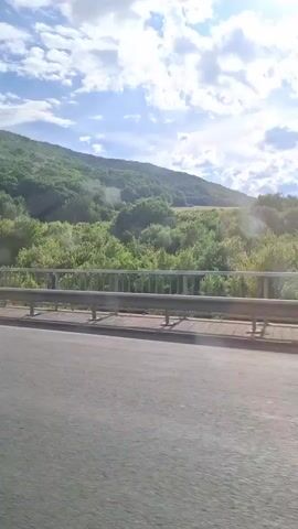 The video shows a view from a bridge overlooking a lush green hillside covered in trees. The sun is shining through white clouds, creating a bright and airy atmosphere. The bridge has a metal railing and a paved road surface is visible in the foreground.