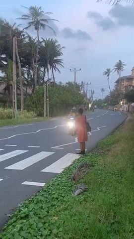 A person stands at a pedestrian crossing on the side of a road. They are wearing a red dress and holding a dark bag over their right shoulder. The person is facing away from the camera and looking towards the road. A motorcycle passes by on the left side of the frame, with its headlight illuminated. The road is paved and has white markings. There are palm trees and other vegetation on the sides of the road. The sky is overcast. In the background, there are more trees, buildings, and several other vehicles on the road, including a tuk-tuk. The overall scene appears to be in a tropical or subtropical location.