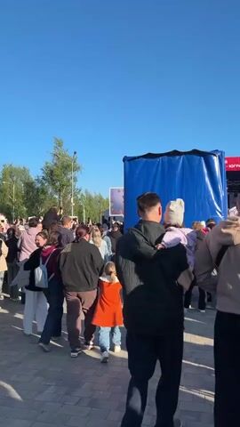 A crowd of people walk across a brick-paved plaza on a sunny day. The camera follows some of them. Some of the people in the crowd are holding or carrying small children. In the distance, a stage is set up with a banner on it. There are tents and a building visible as well. A soundtrack is playing.