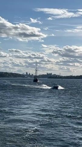 A wide shot of a body of water on a cloudy day. In the foreground, a fast-moving speedboat is creating a large wake. In the mid-ground, a tall-masted sailboat moves at a slower pace. In the background, a city skyline is visible on the horizon. The water is choppy, and the sky is filled with dramatic clouds. The speedboat is white with a dark interior. The sailboat is dark with a single mast. The city skyline is a mix of buildings of varying heights. The overall impression is one of movement and a vast open space.