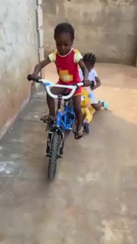 The video shows a young child riding a blue bicycle with training wheels on a concrete surface next to a building. The child wears a red and yellow outfit and sandals, and grips the white handlebars of the bike. A second, slightly younger child follows behind, pushing a yellow toy. The setting appears to be an outdoor courtyard or narrow yard adjacent to the building, which has a textured wall. The child on the bike is concentrating on steering.