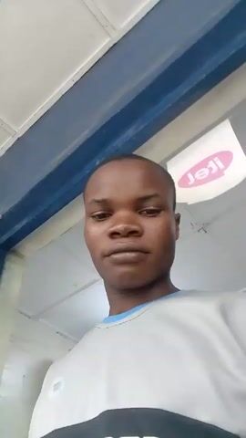 A young, dark-skinned person is recording a video of themself. They are wearing a gray shirt with a black stripe across the chest that says "EASY BUY. BUY NOW PAY LATER." The background shows what appears to be the interior of a shop, with signs visible and light fixtures hanging from the ceiling. The camera angle is slightly low, pointing up at the person.
