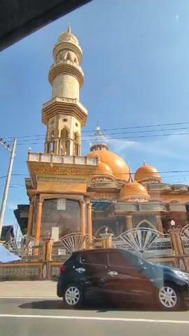 Here's a description of the video:

The video shows a mosque with a tall minaret and several golden domes against a bright blue sky. The mosque is ornate, with intricate architectural details on its exterior. It is surrounded by a decorative fence.

In the foreground, the camera appears to be moving along the street. A person wearing a helmet is riding a white scooter in the same direction as the camera. Later, another person walks by on the sidewalk.  Power lines are visible overhead, stretching across the sky. 

The general impression is that of a sunny day in a location with a prominent Muslim place of worship.